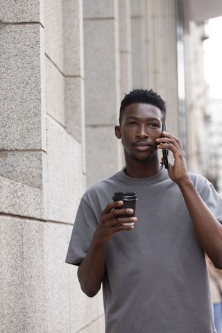 Young man holding coffee and talking on smartphone on urban sidewalk
