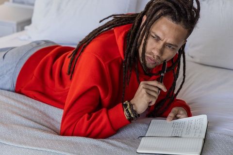 African american man in red hoodie lying on bed writing in notebook planning goals and reflecting