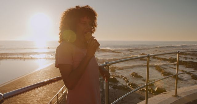 Happy biracial woman eating ice cream standing on promenade by the sea at sundown, copy space. Summer, vacations, lifestyle and free time, unaltered.