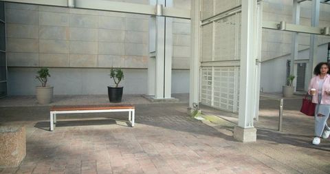 Woman walking through modern urban plaza carrying coffee and red tote bag, sunlit courtyard