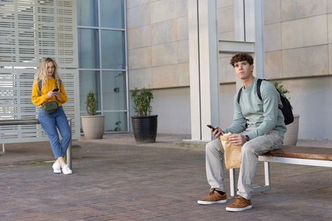 Young adults using smartphones in modern urban courtyard with bench backpacks lunch bag