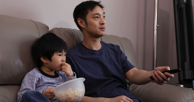 This stock photo captures a father and son enjoying a movie night at home on a comfortable couch with a bowl of popcorn. Ideal for use in parenting blogs, family-oriented advertisements, or articles promoting relaxation and family bonding.