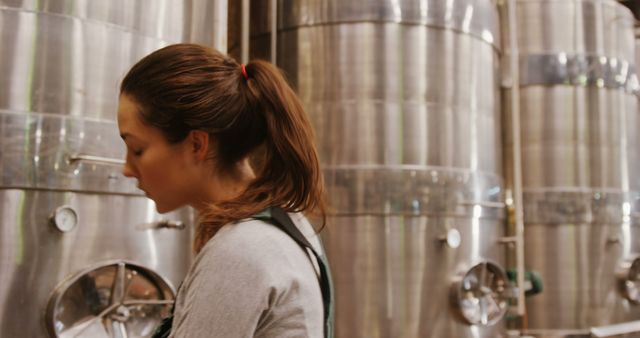 Female worker inspecting large stainless steel fermentation tanks in a microbrewery. Ideal for content related to industrial processes, brewing, manufacturing, and craft beer production. Useful for articles, advertisements, or educational content about the brewing industry and production facilities.