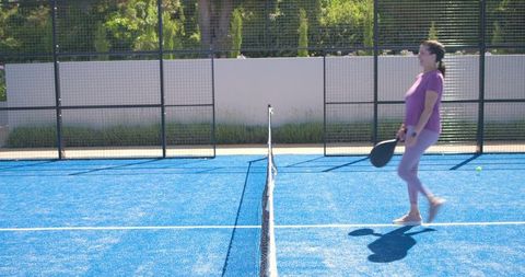 Mature woman in purple activewear holding paddle on blue padel court walking along net