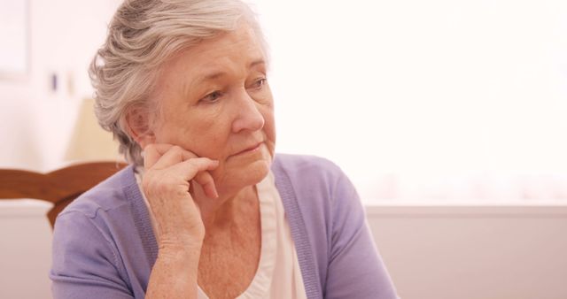 This image features a senior woman sitting indoors, appearing deep in thought. It is a good visual for topics related to aging, elderly care, mental health, contemplation, and a peaceful home environment. It can be used in articles, brochures, or websites focusing on senior living, healthcare, or mental wellness.