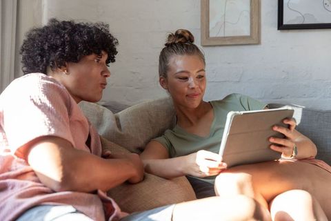 Diverse friends relaxing on sofa sharing tablet in sunlit cozy living room