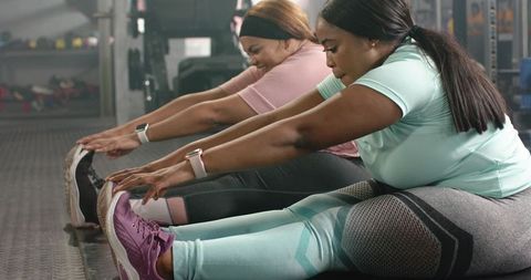 Women stretching together on gym floor reaching toes wearing smartwatches and leggings