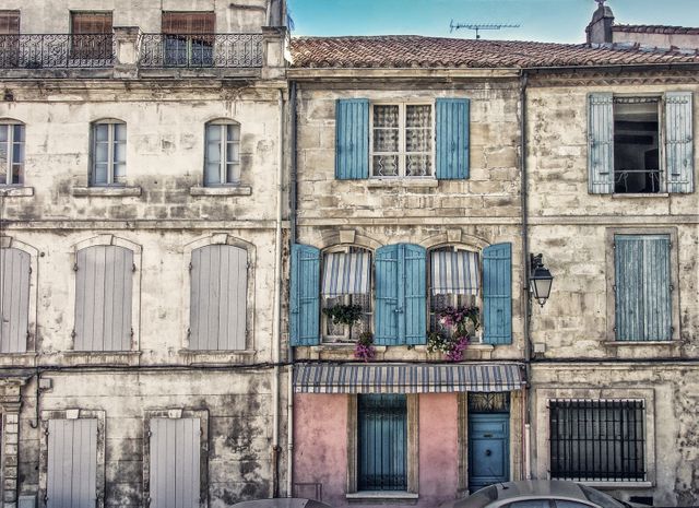 Charming old French building with colorful shutters and traditional architecture. Blue and pink shutters add a pop of color to the historic stone facade. Windows with floral boxes enhance the scenic beauty. Ideal for travel blogs, architectural design inspiration, historic preservation topics, and European cultural exposure materials.