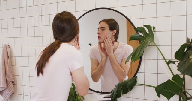 Young woman with long hair checking her skin in round bathroom mirror, focusing on skin care routine. Green plants and white tiled wall visible in background. Suitable for promoting beauty routines, skin care products, self-care practices, or wellness blogs.