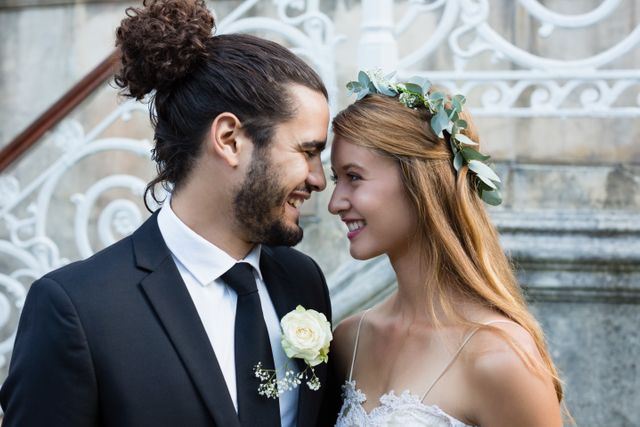 This image captures a joyful moment between a bride and groom standing face to face, smiling at each other. The bride is wearing a floral crown and a lace dress, while the groom is dressed in a formal suit with a boutonniere. Ideal for use in wedding invitations, romantic greeting cards, wedding blogs, and articles about wedding planning and love stories.