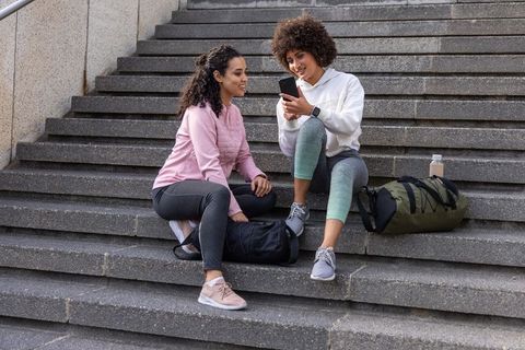 Friends sitting on city steps sharing smartphone after workout session with duffel bags