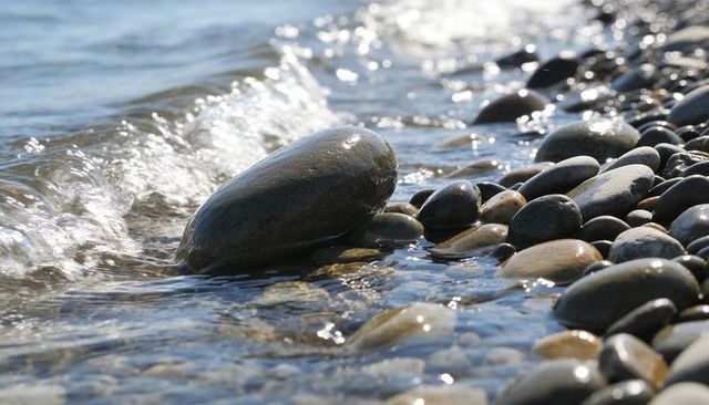 Glistening smooth stone on pebble shore with lapping wave and sunlit reflections