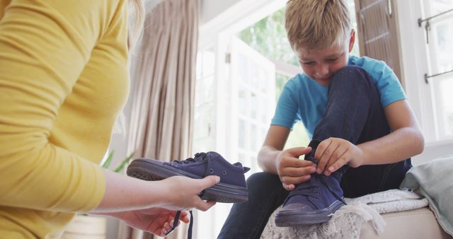 Young boy receiving help from his mother to tie his shoes at home, portraying the healthy caregiver-child relationship. This moment captures family bonding, skill learning, and home comfort, making it suitable for parenting blogs, family lifestyle articles, advertisements related to children's apparel, or guides on teaching self-care skills to children.