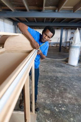 African american carpenter measuring and marking cabinet frame in woodworking workshop