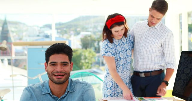 Man smiling confidently at camera while two coworkers discuss a project in the background. Provides a vibrant depiction of teamwork and a collaborative office culture. Ideal for business-related content, workplace environment themes, corporate training materials, or marketing campaigns focusing on teamwork and professionalism.