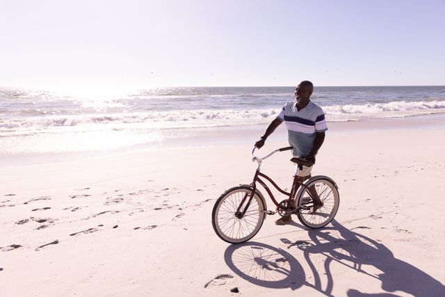 African american senior man with bicycle walking at sandy beach against sea and sky on sunny day. Copy space, travel, retirement, unaltered, lifestyle, vacation, scenic, enjoyment and nature concept.