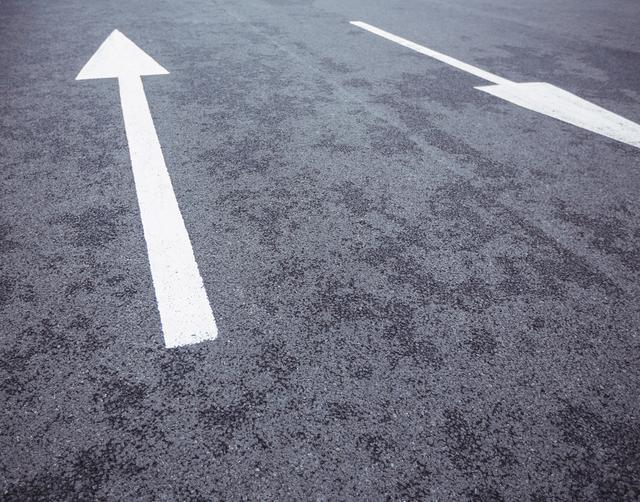 Image shows two white arrows on an asphalt road pointing in opposite directions. Useful for illustrating concepts of direction, traffic flow, navigation, and urban transportation planning.