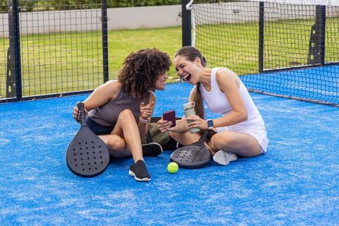 Laughing friends sitting on blue padel court holding paddles and checking phone