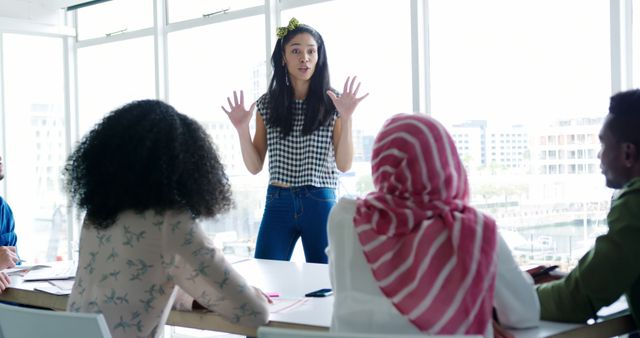 Young woman wearing casual attire actively presenting ideas in front of diverse team in modern open office with large glass windows during daytime. Suitable for concepts of teamwork, collaboration, business discussions, brainstorming sessions, office dynamics, and modern work environments.