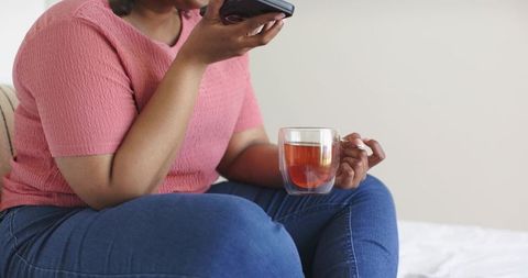 African american woman holding glass mug of tea while using smartphone in cozy bedroom