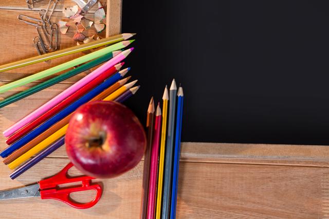 Colorful school supplies including colored pencils, an apple, scissors, paper clips, and pencil shavings on a wooden desk with a chalkboard. Ideal for educational content, back-to-school promotions, classroom decor, and creative learning materials.