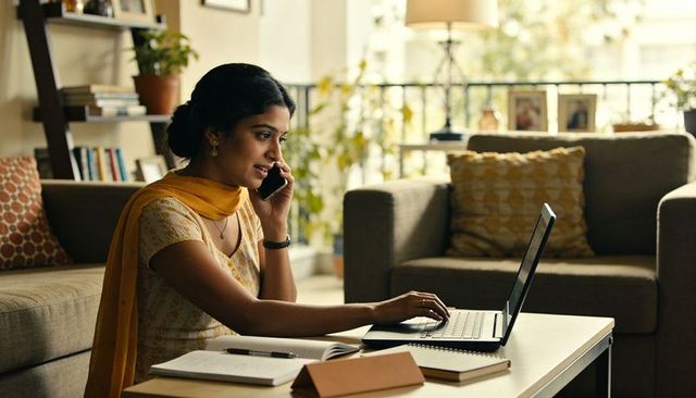 Indian woman remote working calling on smartphone while typing on laptop in living room