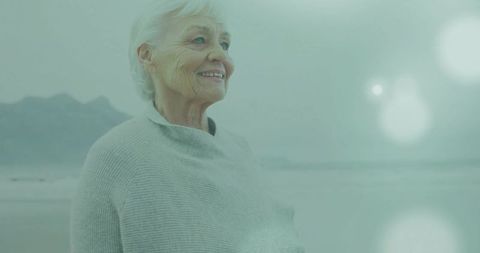 Senior woman standing on misty beach smiling, contemplative seaside wellness lifestyle