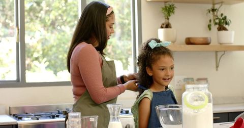 Hispanic mother smoothing daughter hair while baking together in sunlit family kitchen