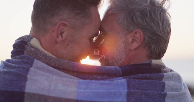 Happy caucasian gay male couple wearing blanket, embracing and taking selfie at sunset on the beach. summer road trip and holiday in nature.
