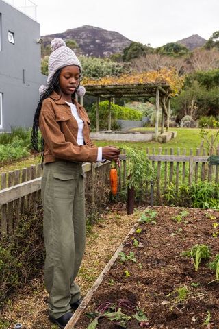 Teen african american girl harvesting carrot in backyard garden wearing knit beanie