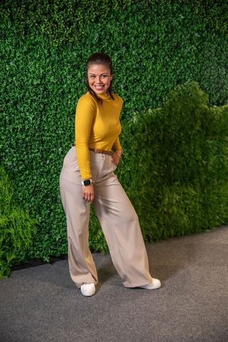 Confident woman posing and smiling by green wall wearing mustard top and beige trousers
