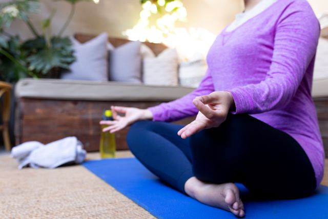 Low section of biracial woman sitting in yoga meditation pose on mat in living room, copy space. Happiness, health, fitness, inclusivity and domestic life.