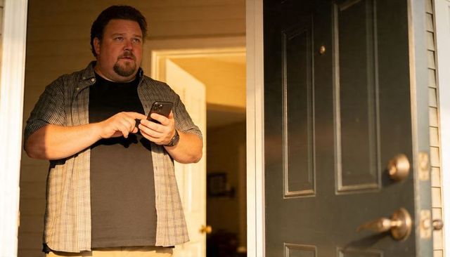 Middle-aged man standing at front doorway holding smartphone and checking screen at dusk