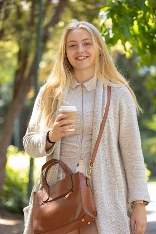 Young woman smiling holding takeaway coffee walking sunlit park with leather handbag