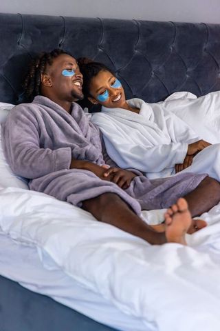 Relaxing couple wearing blue under-eye patches in plush robes, lounging on tufted headboard