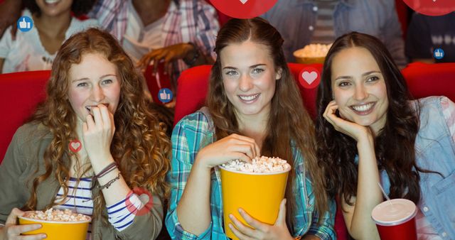 Three young women sitting in a movie theater, enjoying the movie with popcorn. Social media reaction icons appear around them, indicating an engaging and social experience. Useful for concepts related to entertainment, social engagement, friendship, and leisure activities.