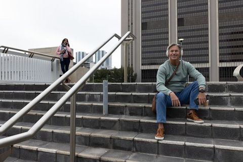 Mature man sitting on city steps listening with headphones holding coffee and phone
