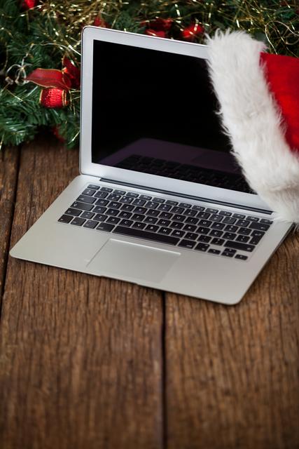 This image features a laptop adorned with a Santa hat, placed on a wooden plank, surrounded by festive Christmas decorations. It is perfect for holiday-themed marketing materials, blog posts about technology during the holiday season, or social media posts celebrating Christmas and New Year. The combination of technology and festive elements makes it ideal for promoting holiday sales, digital greetings, or seasonal offers.
