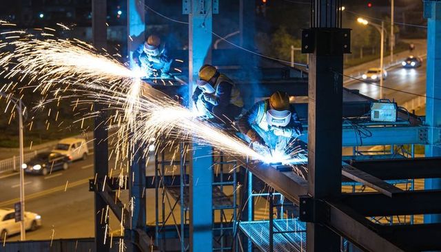 Night welding crew sparking steel beams over busy urban roadway, high-rise construction