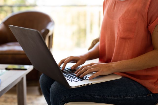 Caucasian woman sitting on a couch in her living room, typing on a laptop. Her phone is on the table in the background. This image can be used for themes related to remote work, modern lifestyle, technology, and casual home settings.