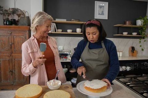 Mature friends frosting layer cake together in cozy kitchen sharing baking moment