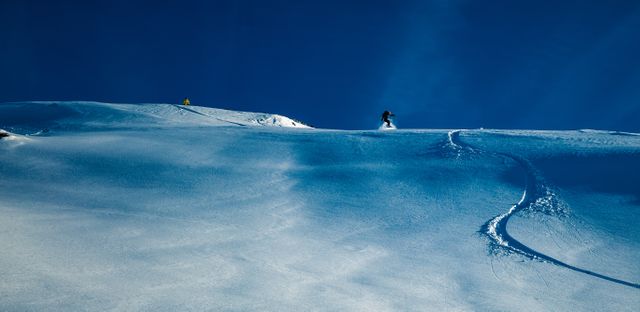 Snowboarders are gliding down a pristine snowy mountain under a clear blue sky. This stock photo captures the thrill of snowboarding in an untouched winter landscape, ideal for winter sports advertising, travel brochures, adventure blogs, or nature magazines. Perfect for conveying excitement, recreational activities, and the beauty of alpine environments.