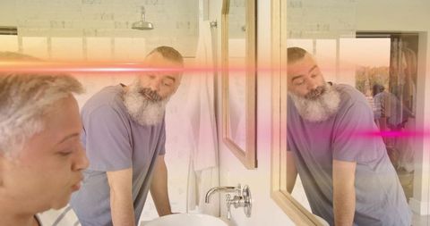 Bearded man leaning at bathroom vanity looking in mirror while partner brushing teeth