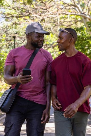 African american friends walking and talking in sunlit park canopy with smartphone