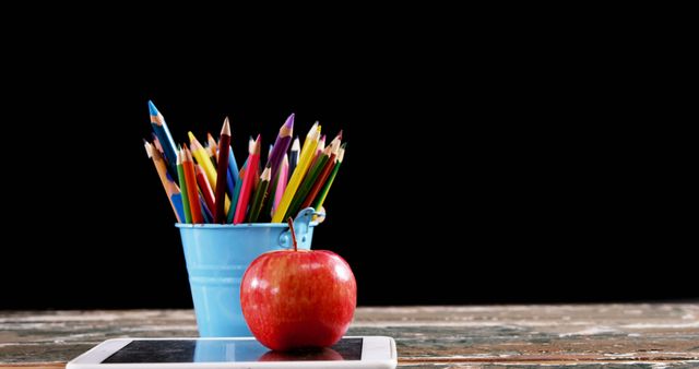 Bright pencils in blue holder along with red apple on wooden table symbolize education and creativity. Ideal for use in education themes, back-to-school promotions, classroom decorations, study habit illustrations, and school supply advertisements.