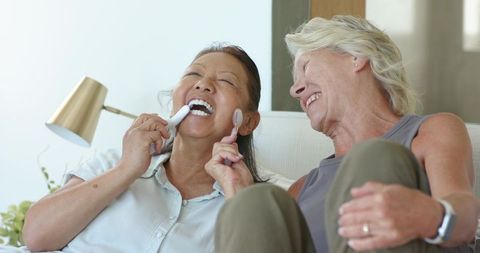 Senior women laughing while checking teeth with toothbrush and mirror on cozy sofa