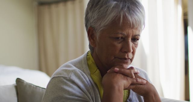 Senior woman with gray hair contemplating indoors, sitting near window with natural light streaming in. Ideal for use in articles about aging, retirement, mental health, and loneliness. Suitable for healthcare and wellness publications, blogs, and social media content emphasizing introspection and thoughtful moments.