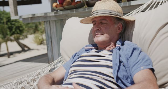 Mature man wearing a straw hat and a striped shirt is relaxing in a hammock at a beachside resort on a sunny day. Ideal for representing relaxation, retirement, summer vacation, or peaceful outdoor leisure. Useful for travel brochures, lifestyle blogs, wellness retreats, and marketing materials.
