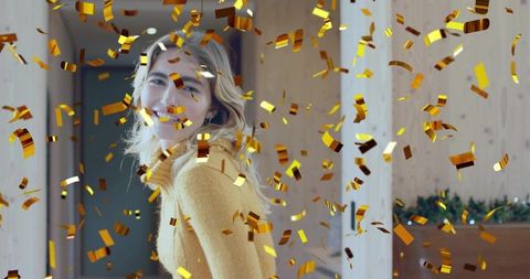 Smiling woman turning in cozy wood hallway celebrating with gold confetti wearing mustard sweater
