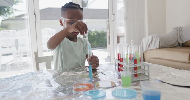 Young African American boy wearing safety goggles conducting science experiments at home. He is mixing chemicals in test tubes on a table covered with foil, showcasing curiosity and enthusiasm for science. This can be used for educational content, homeschooling materials, or promoting STEM education for children.